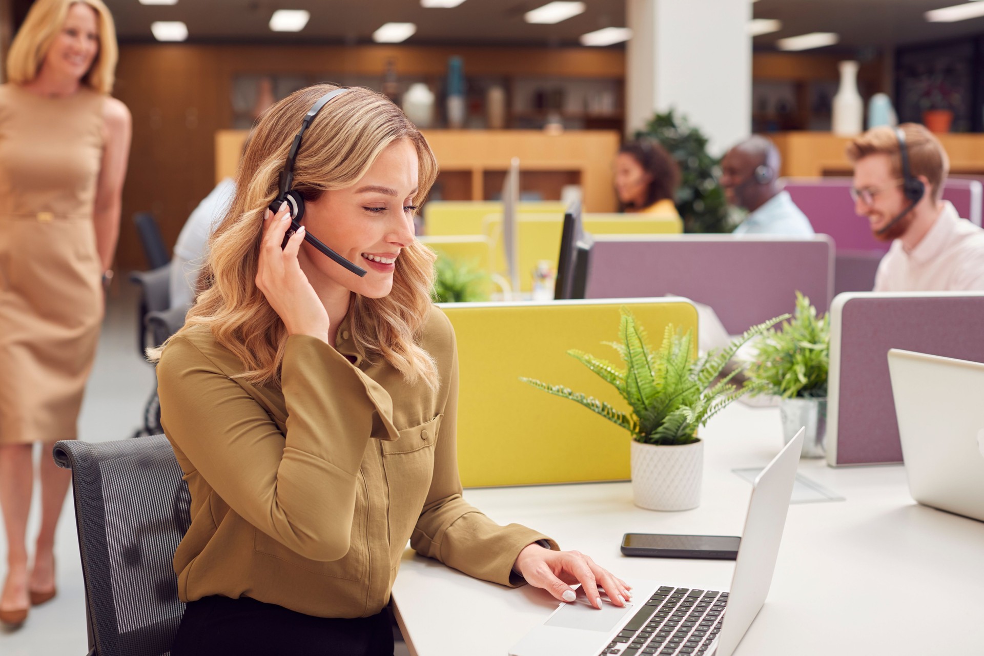 Businesswoman Wearing Phone Headset Talking To Caller In Busy Customer Services Centre